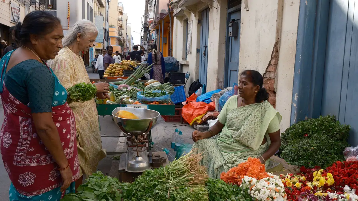 Women buy greens and flowers from a vendor seated beside her produce on a market street