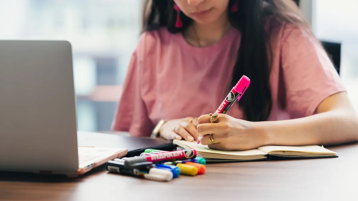 A woman in a pink T-shirt writes in a notebook with a pink highlighter beside a laptop and a pile of coloured markers