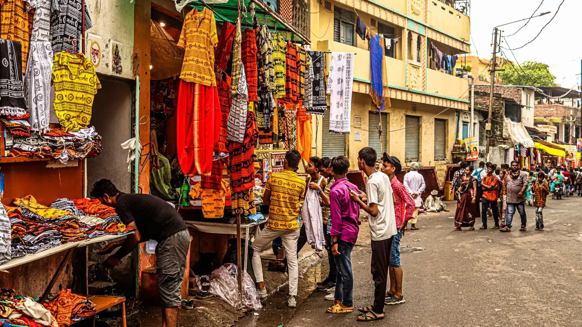 Shoppers browse clothing stalls hung with colourful printed shirts and fabrics along a busy market street in India