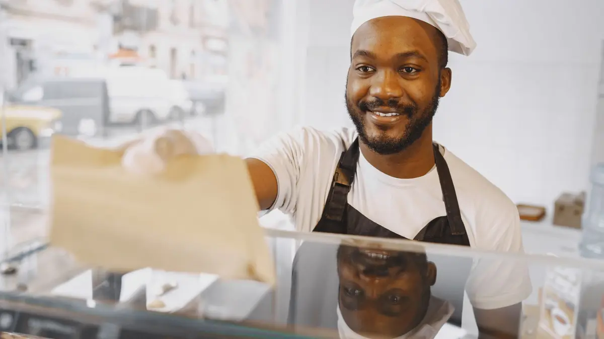 A smiling baker in a cap and apron hands a paper bag across the counter to a customer