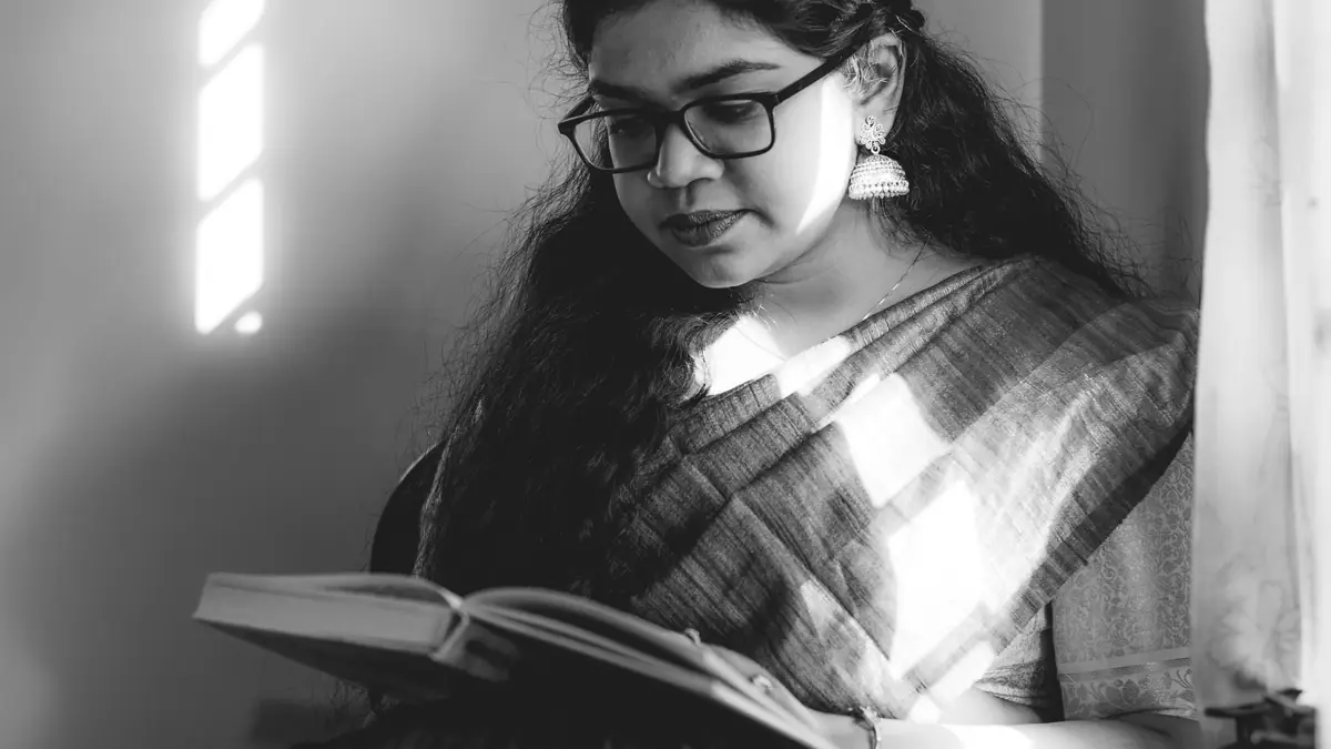 A woman in glasses and a sari reads a book by window light, in black and white