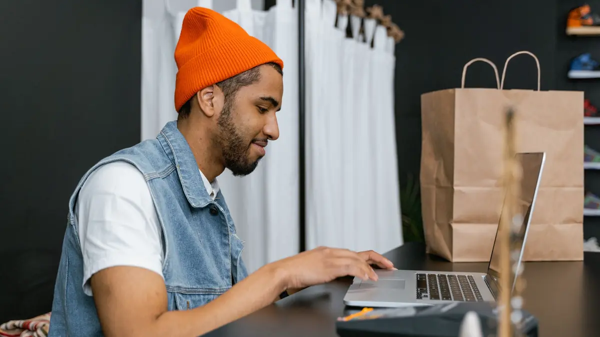A shop owner in an orange beanie works on a laptop at his counter, with a paper shopping bag beside him