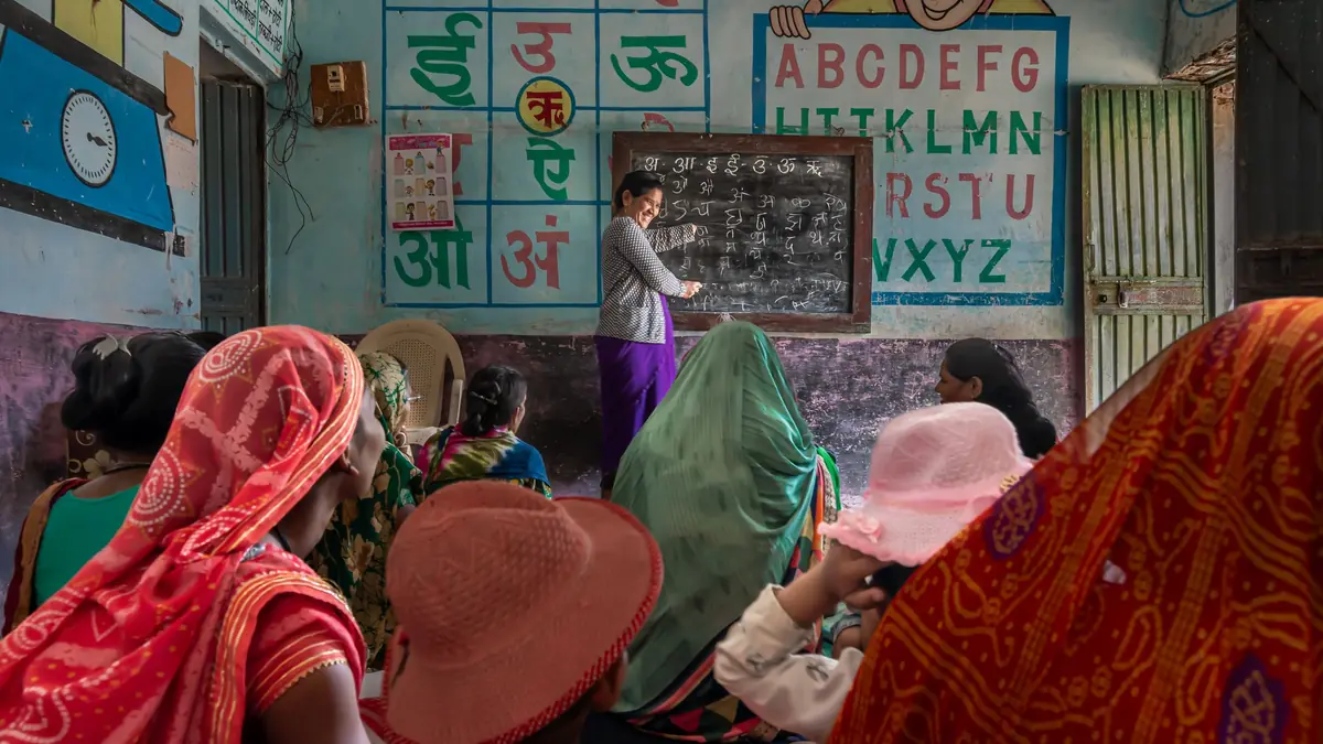 A teacher writes Hindi letters on a blackboard for a class of women seated on the floor, with alphabet charts painted on the wall