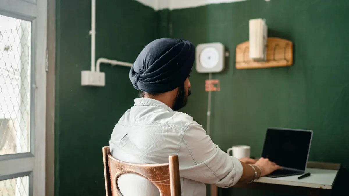 A man in a turban, seen from behind, works on a laptop at a small desk by a window