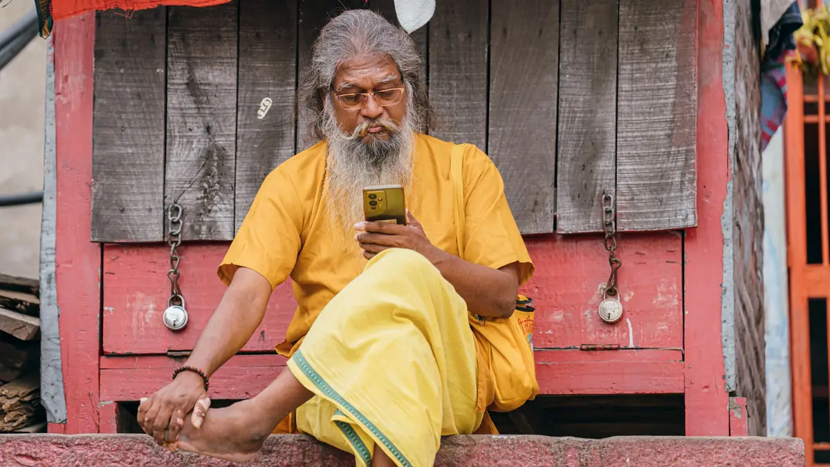 An older man with a white beard, in a yellow kurta and dhoti, looks at his smartphone on a doorstep in Varanasi