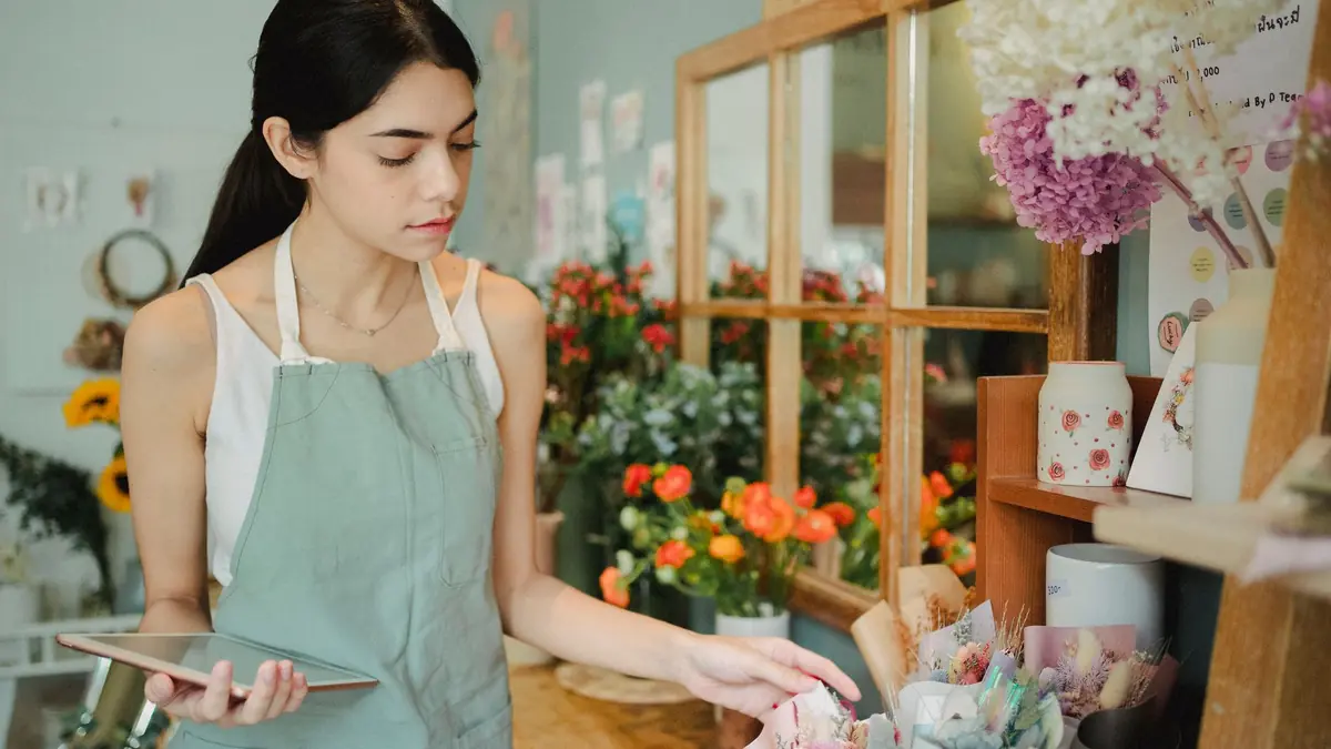 A young woman in an apron holds a tablet while arranging flowers at the counter of her flower shop