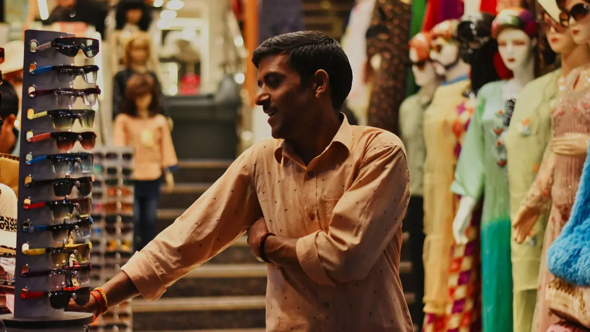 A smiling shopkeeper leans on a sunglasses stand in a clothes shop with outfits on mannequins