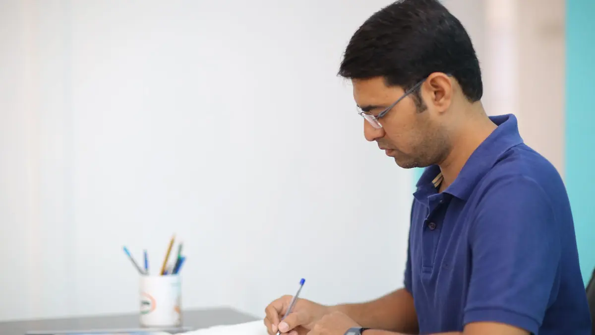 A man in a blue polo shirt and glasses writes in a notebook at a white desk